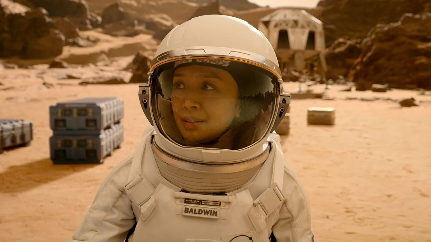 Nahaufnahme einer Astronautin auf dem Mars. Im Hintergrund sind mehrere Container und ein weißes Gebäude zu sehen.