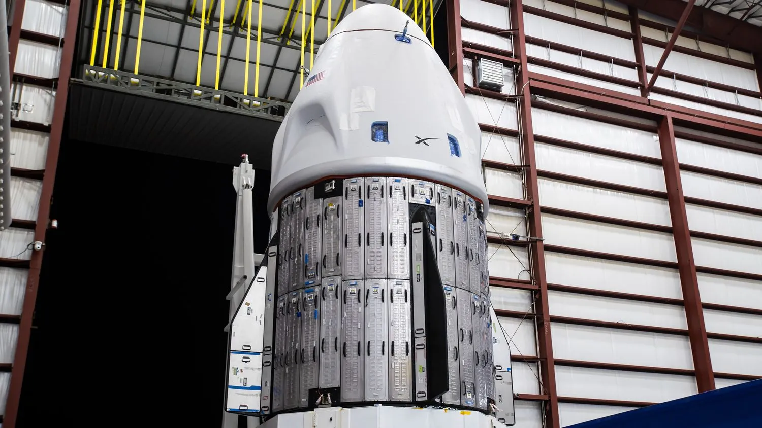 eine weiße SpaceX-Dragon-Kapsel ist in einem großen Hangar in der Nähe einer Tür zu sehen, die in die Dunkelheit führt.