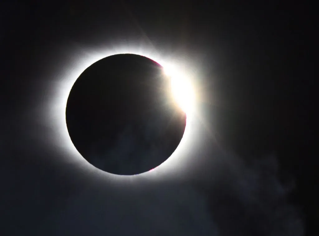 Foto einer Sonnenfinsternis, auf dem ein weißer Umriss um einen zentralen schwarzen Kreis zu sehen ist, hinter dem in der rechten oberen Ecke ein helles Licht leuchtet.