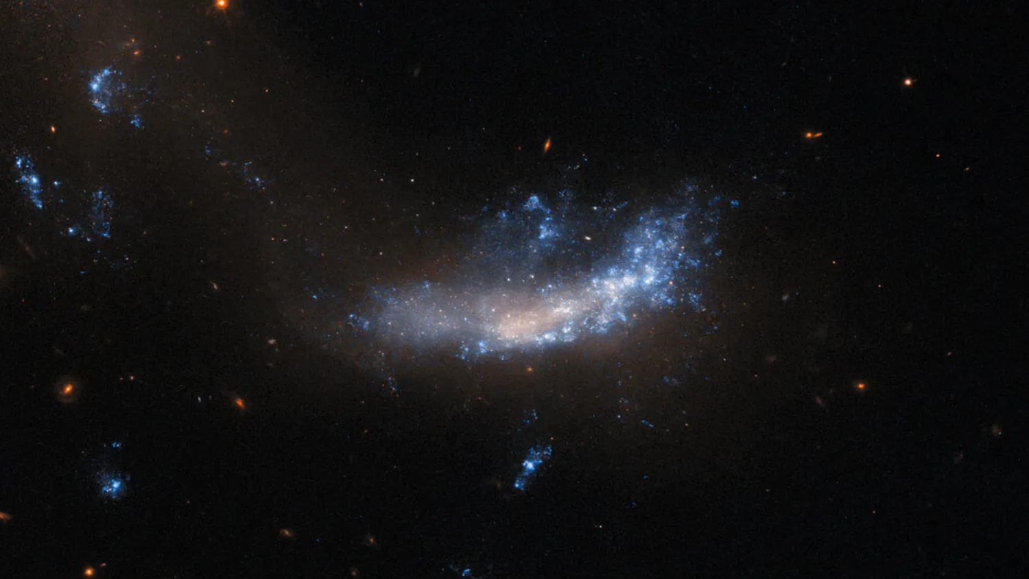 Eine Galaxie, die flach und unförmig ist. Oben und auf der rechten Seite ist sie von leuchtenden Gas- und Staubfahnen bedeckt, während ihr Zentrum und ihre linke Seite eher trübe und fleckig sind. Eine Spur aus dunklem, trübem Staub zieht sich von unterhalb der Galaxie nach oben und nach links, wo sich drei weitere helle Flecken befinden. Der Hintergrund um die Galaxie ist recht dunkel, nur ein paar kleine Hintergrundgalaxien und ein Stern sind zu sehen