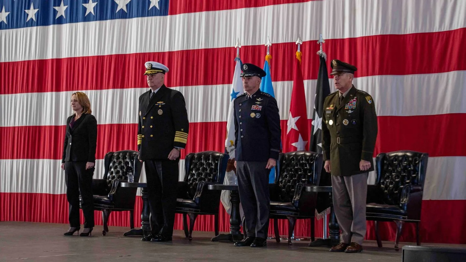 vier Personen, drei davon in US-Militäruniformen, stehen vor einer amerikanischen Flagge