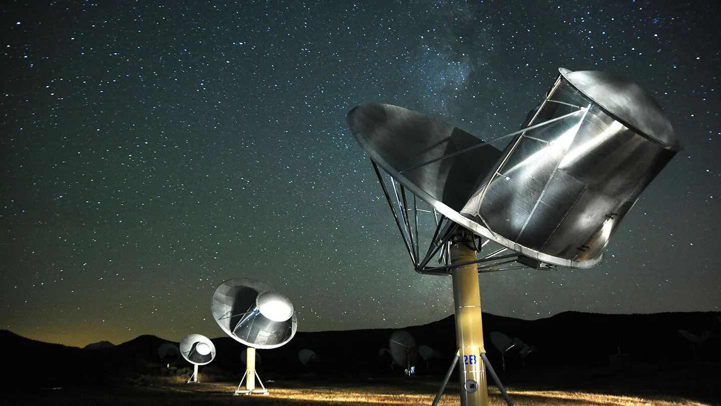 Punktbeleuchtete Satellitenschüsseln in einer kargen Landschaft, die vor dem Hintergrund einer sternenklaren Nacht in den Himmel ragen