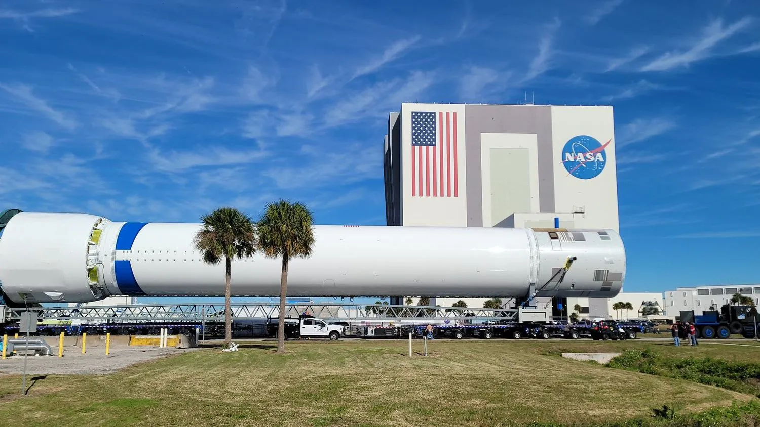 eine große weiße Rakete rollt eine Straße hinunter, mit einem großen NASA-Gebäude und blauem Himmel im Hintergrund.