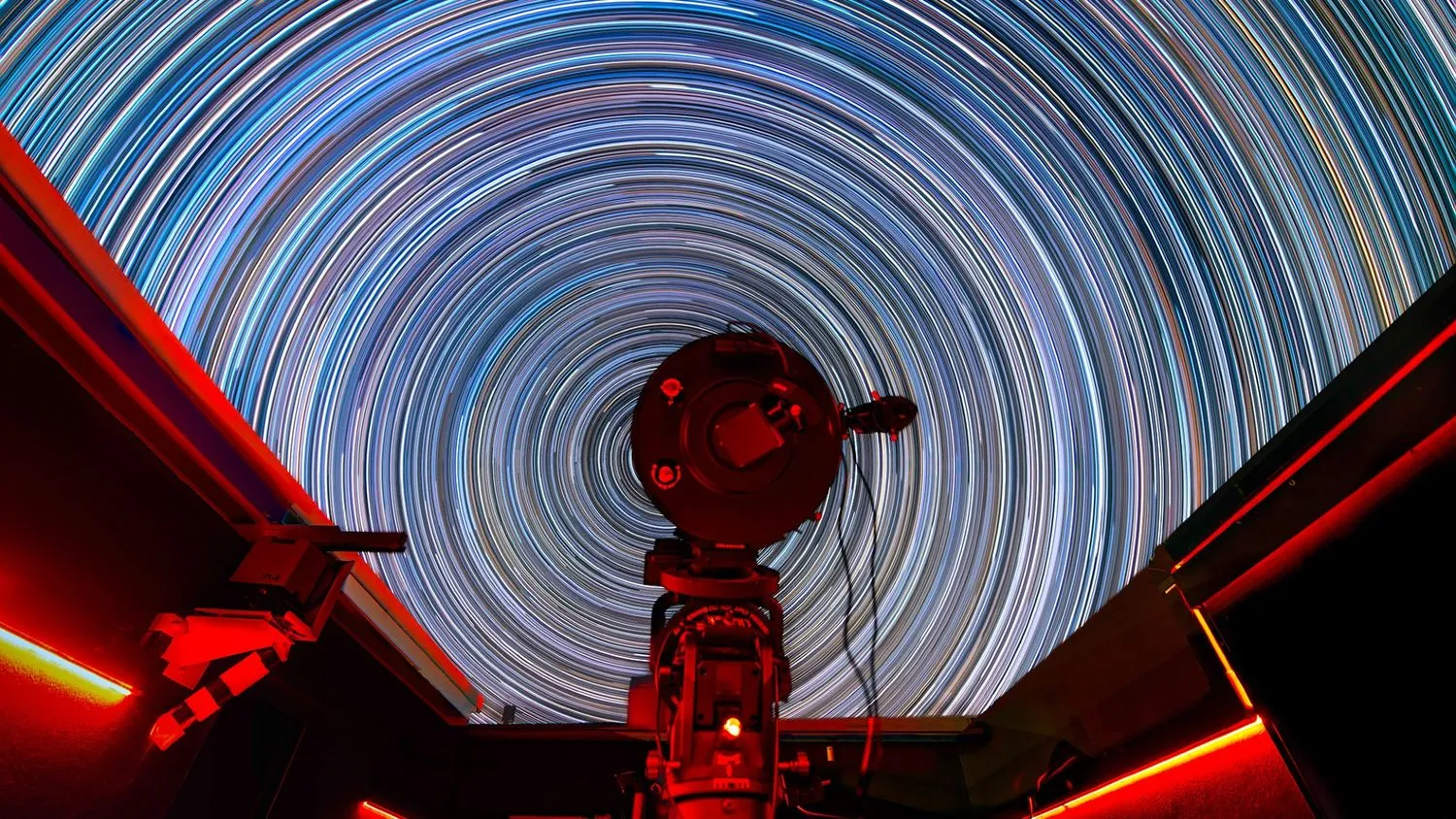 im vordergrund ist ein teleskop zu sehen. im hintergrund verwandeln sich die sterne dank einer langzeitbelichtung in riesige ringe mit verschiedenen farben am himmel