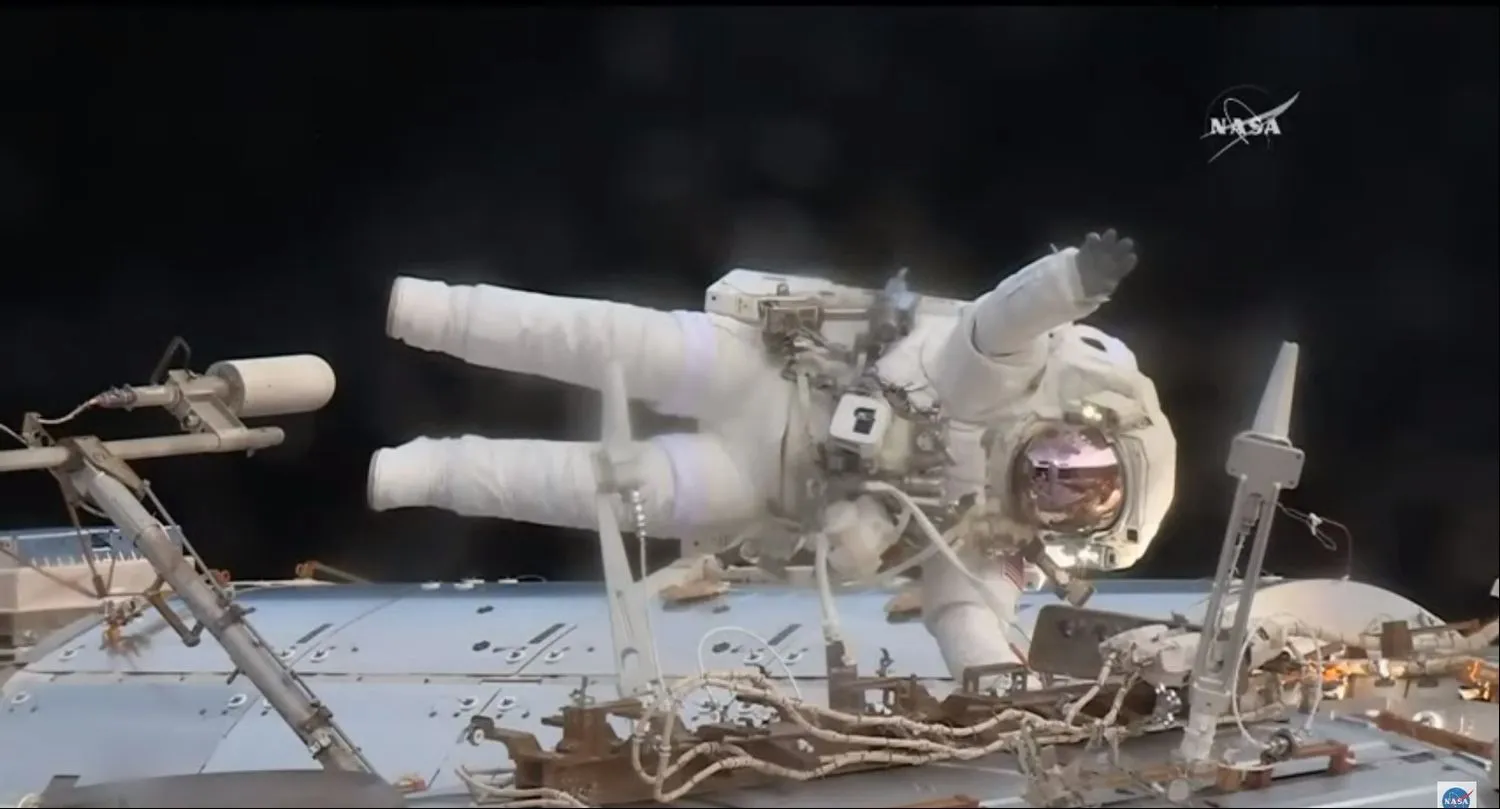 ein astronaut im raumanzug schwebt über einem modul. er hält sich mit einer hand fest und winkt mit der anderen. aus dem aufnahmewinkel liegt er auf der seite