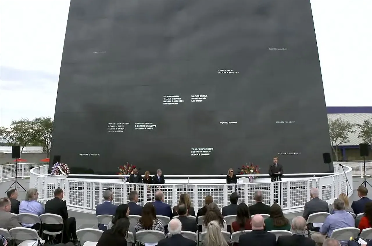 eine Handvoll Menschen in dunklen Anzügen steht vor einer großen schwarzen Wand, in die mehrere Dutzend Namen eingemeißelt sind.