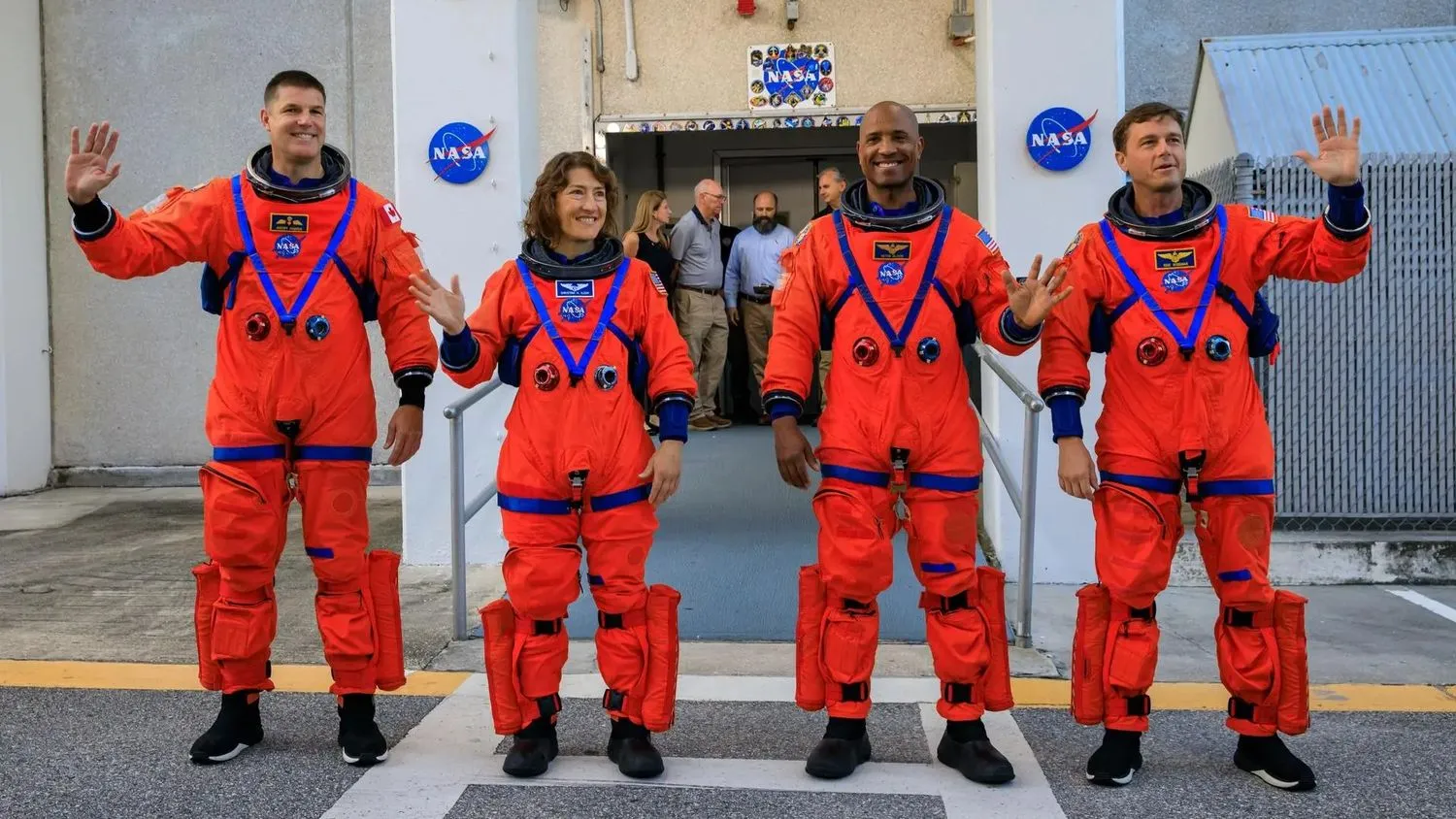 vier astronauten in orangefarbenen raumanzügen winken vor einem mit nasa-logos geschmückten eingang in die kamera