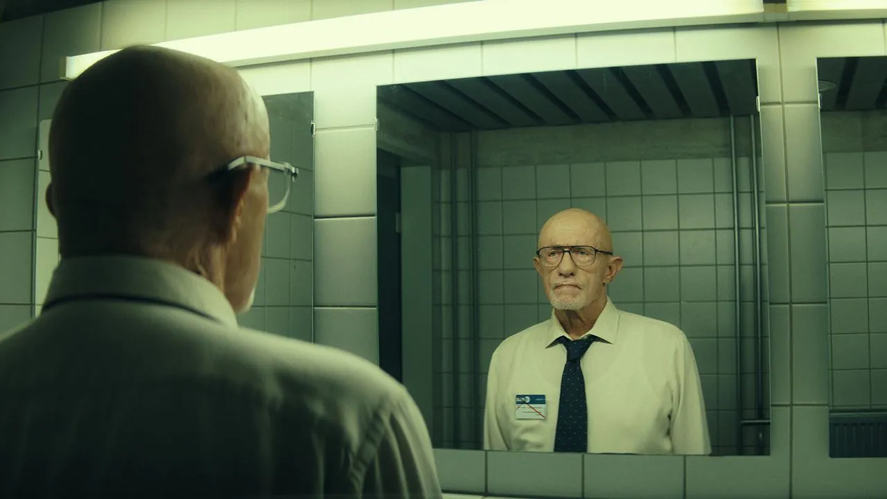 Ein glatzköpfiger Mann mit Brille, weißem Hemd und schwarzer Krawatte schaut aufmerksam in den Spiegel einer öffentlichen Toilette.