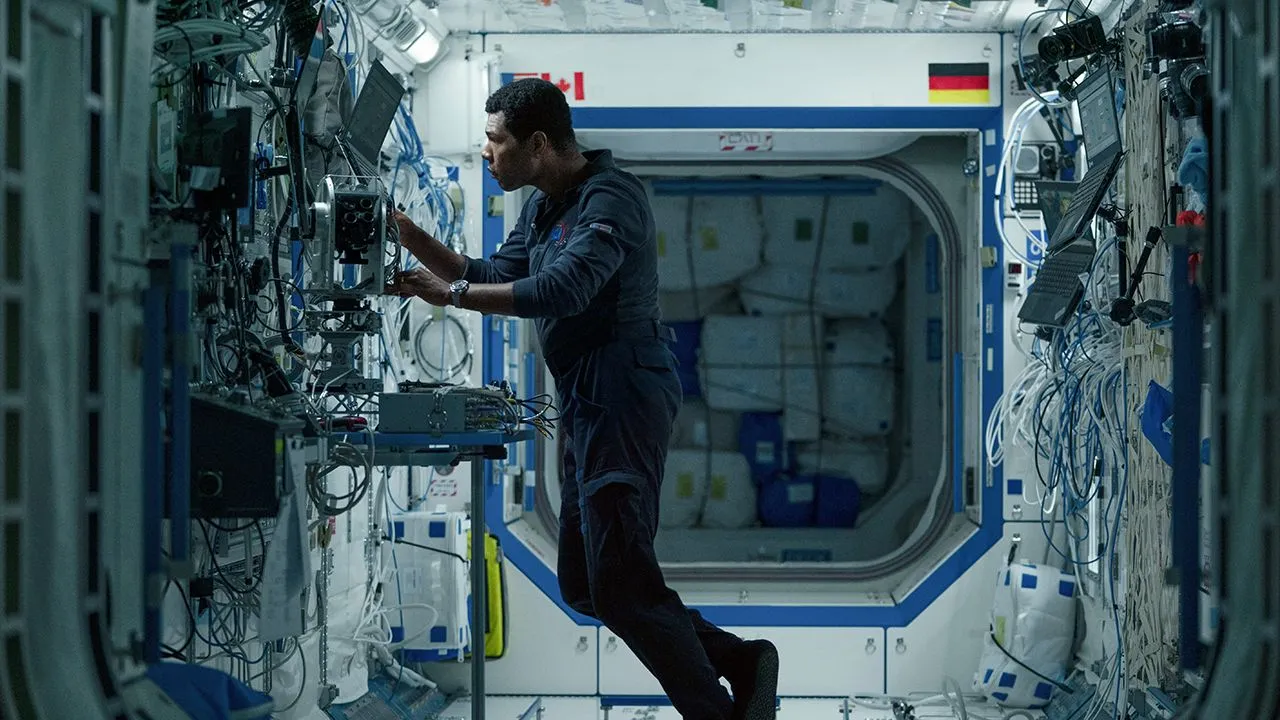 Ein Mann in einem blauen Fluganzug steht in einem beengten Labor voller Drähte und Bildschirme im Weltraum. ER arbeitet an einem der Bildschirme.