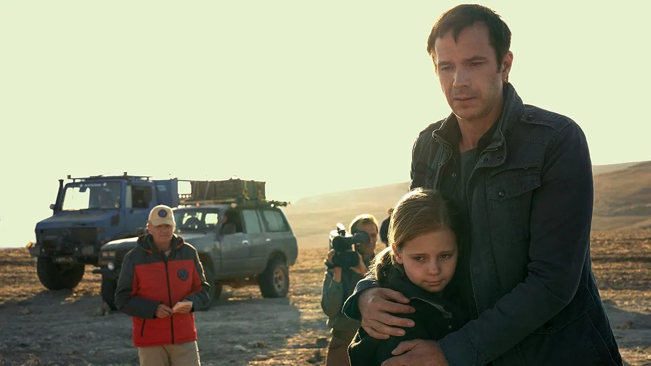 Ein Mann umarmt seine kleine Tochter. Im Hintergrund sind zwei Männer zu sehen, von denen einer eine Kamera in der Hand hält. Außerdem sind zwei große Fahrzeuge zu sehen. Sie befinden sich draußen in einer verlassenen, kargen Landschaft.