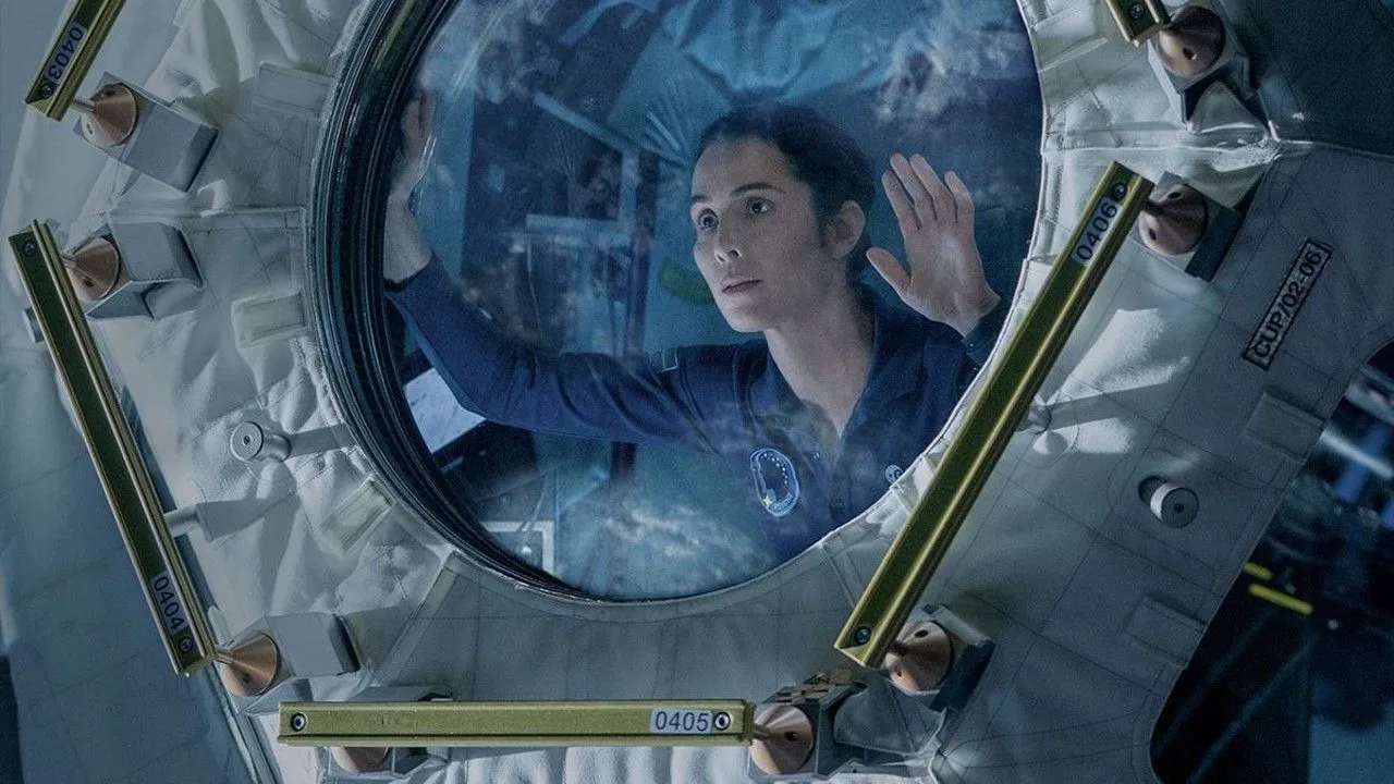 eine Frau in einem blauen Fluganzug schaut durch eine gläserne Luke in einem beengten Labor voller Kabel und Bildschirme