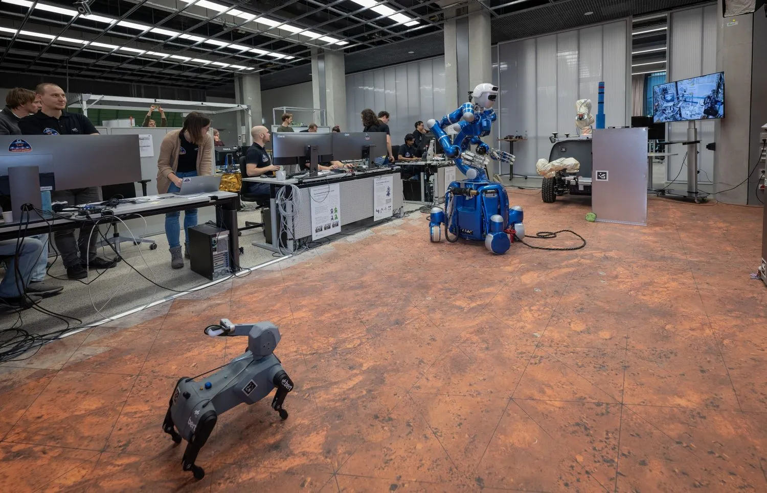 ein hundeähnlicher Roboter und ein humanoider Roboter auf Rädern rollen durch ein Labor, umgeben von Wissenschaftlern in T-Shirts, die an Computern stehen