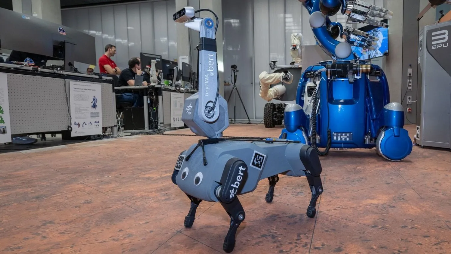 ein vierbeiniger hundeförmiger Roboter mit Kulleraugen und einer dicken Antenne, die als Kopf dient, in einem Labor mit Geräten und Computern