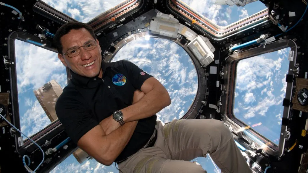 Ein Mann in Khakis verschränkt die Arme, während er vor einem sechseckigen Fenster schwebt, das vom Orbit aus auf die Erde hinunterblickt. 