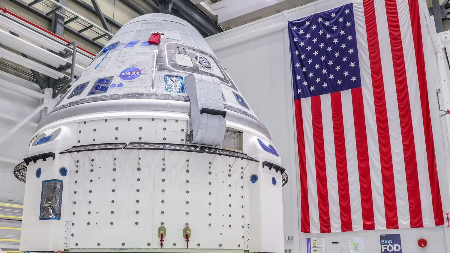 eine silber-weiße Raumkapsel befindet sich in einem riesigen weißwandigen Raum mit einer amerikanischen Flagge im Hintergrund.