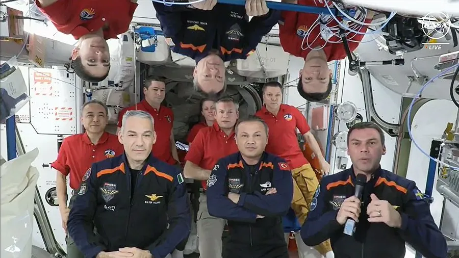 11 Astronauten, 7 in roten Hemden und 4 in blauen und orangenen Fluganzügen auf der Raumstation. einige stehen auf dem Kopf