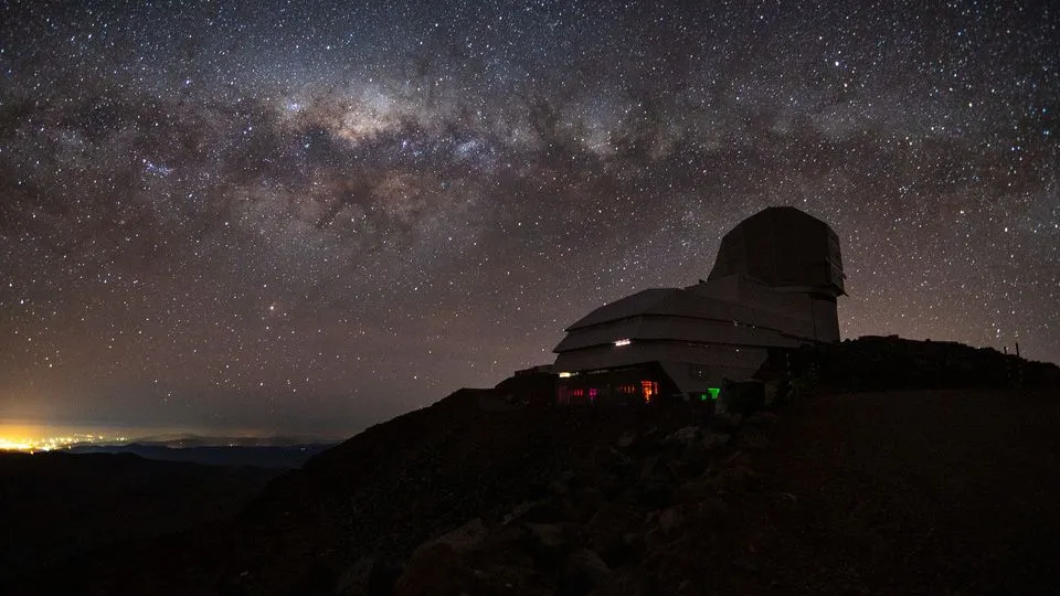 Die Silhouette eines Observatoriums sitzt auf einem dunklen Hügel unter der Milchstraße, die sich über den Sternenhimmel erstreckt.