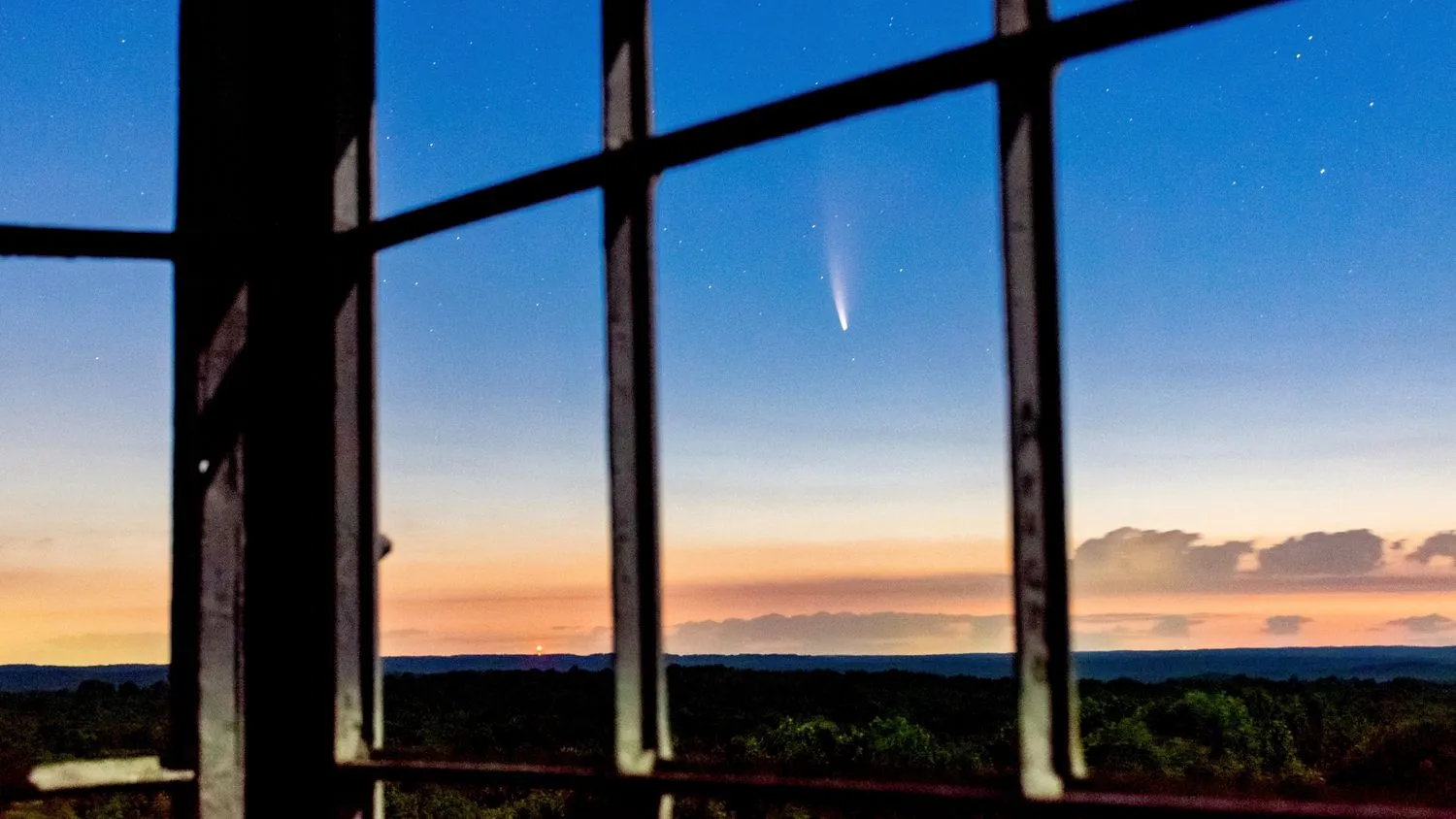 ein Komet streift den Himmel über einem dunklen Wald, gesehen durch leere Fenster hoch oben