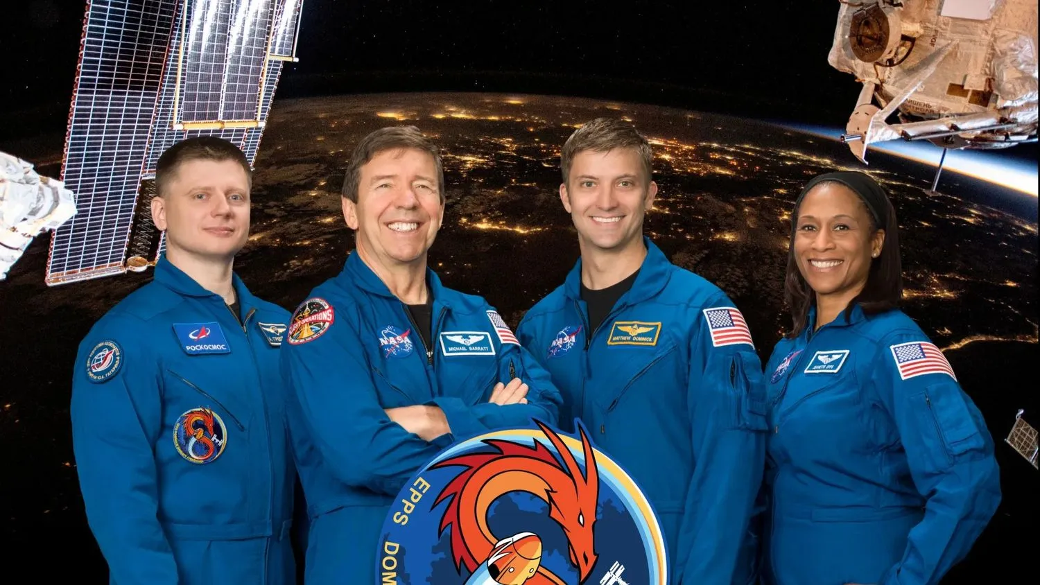 Vier lächelnde Menschen - drei Männer und eine Frau - in blauen Fluganzügen stehen vor einem Bild der nächtlichen Erde, das aus einer niedrigen Erdumlaufbahn aufgenommen wurde.