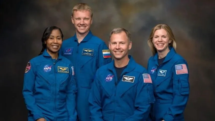 Vier lächelnde Menschen in blauen Fluganzügen stehen vor einem dunkelgrau-braunen Hintergrund.