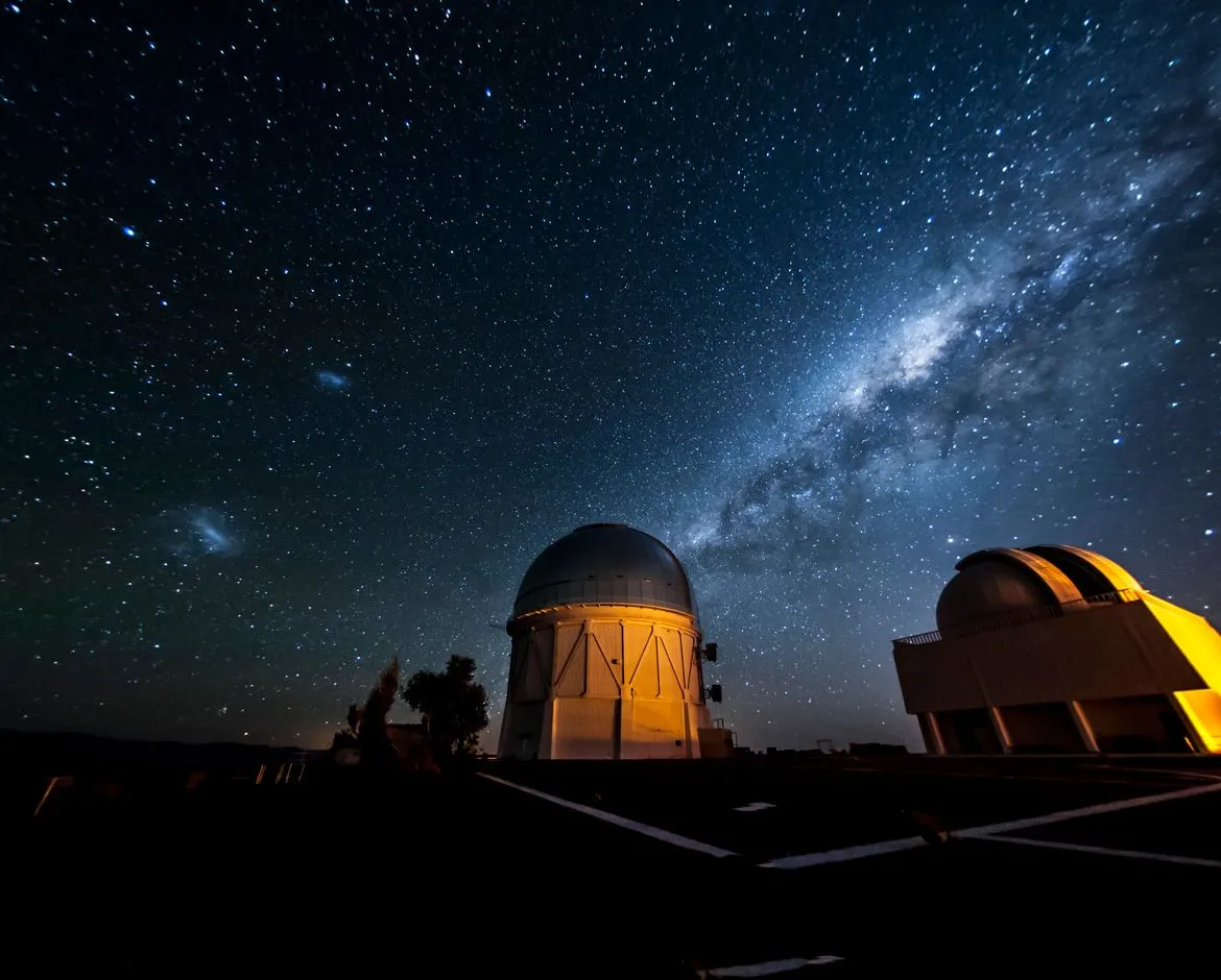 Zwei schwach beleuchtete Observatorien sitzen wie verkümmerte Kuppeln unter einem leuchtenden Nachthimmel und einem Stück blau gefärbter Milchstraße.