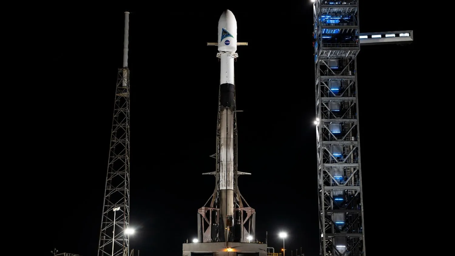 eine Rakete steht aufrecht auf der Startrampe bei Nacht mit einer großen weißen Nutzlast oben drauf