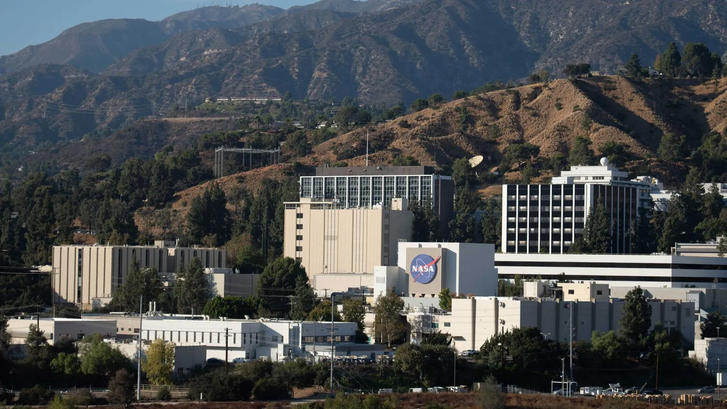eine Handvoll weißer und hellbrauner Gebäude, eines davon mit einem großen blauen NASA-Logo, stehen am Fuße der Berge.