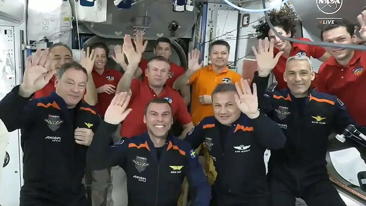 11 Menschen in Fluganzügen winken an Bord der weißwandigen internationalen Raumstation in die Kamera.