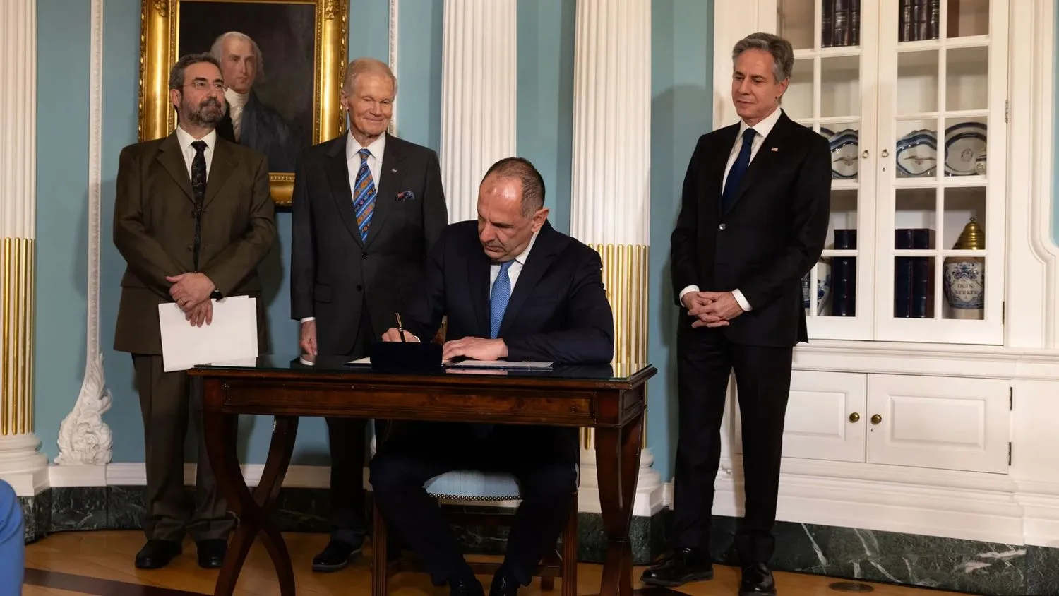 Drei lächelnde Männer in Anzügen stehen hinter einem Mann, der an einem kleinen Holztisch sitzt und ein Dokument unterschreibt.