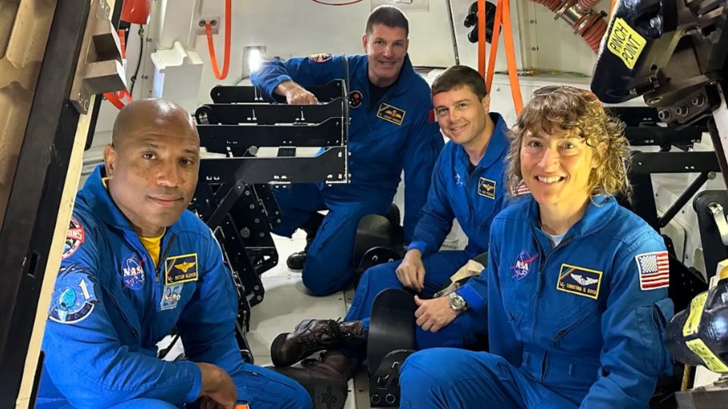 vier Astronauten in einem kleinen Raumschiff, die in Fluganzügen hocken und in die Kamera lächeln
