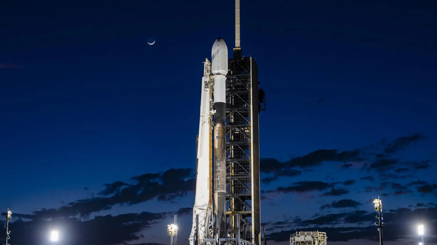 Eine weiß-schwarze SpaceX-Rakete mit der Intuitive Machines IM-1-Mondlandefähre an Bord auf der Startrampe mit einer Mondsichel über dem Kopf.