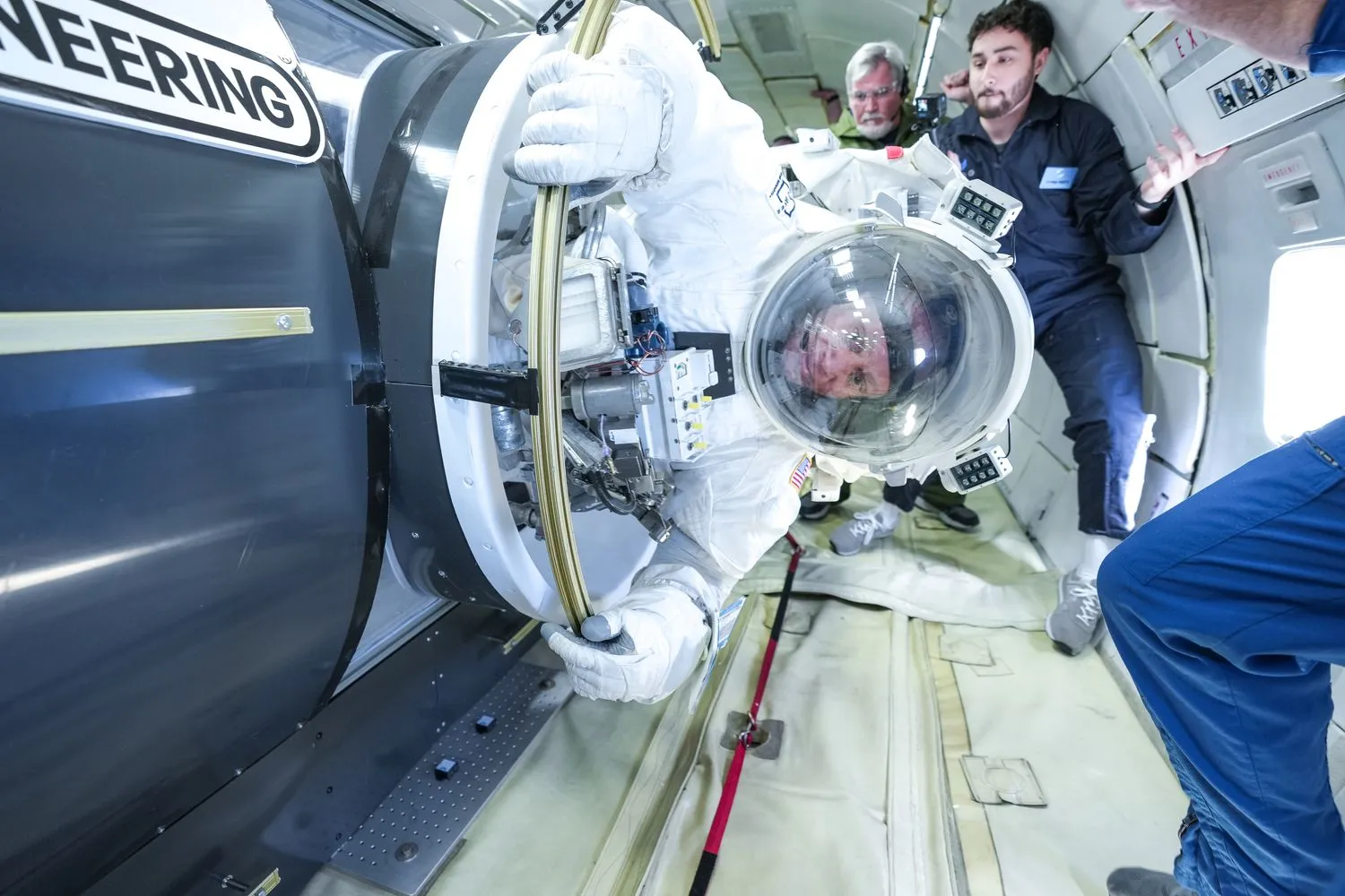 eine Person in einem klobigen Raumanzug schwebt in einem Flugzeugrumpf, umgeben von Menschen in blauen Fluganzügen