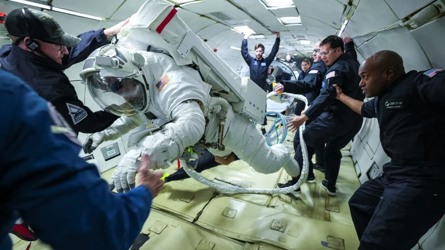 eine Person in einem klobigen Raumanzug schwebt schwerelos im Rumpf eines Flugzeugs, umgeben von Menschen in blauen Fluganzügen und Schutzbrillen