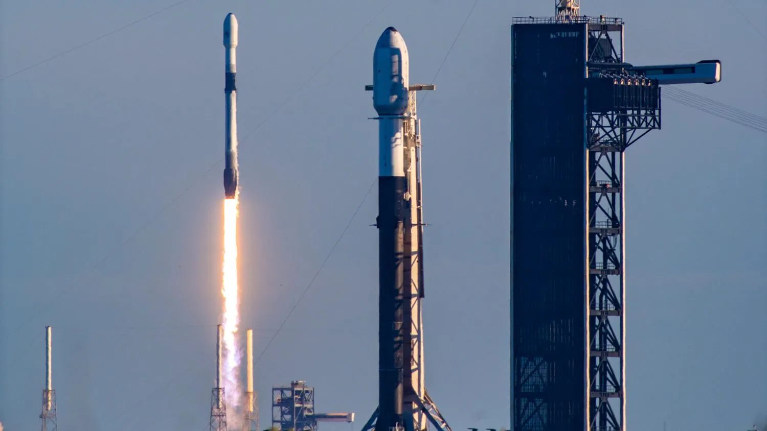 eine weiß-schwarze Spacex Falcon 9 Rakete startet in den blauen Himmel hinter einer anderen Falcon 9, die auf einer Startrampe zum Abheben bereit steht
