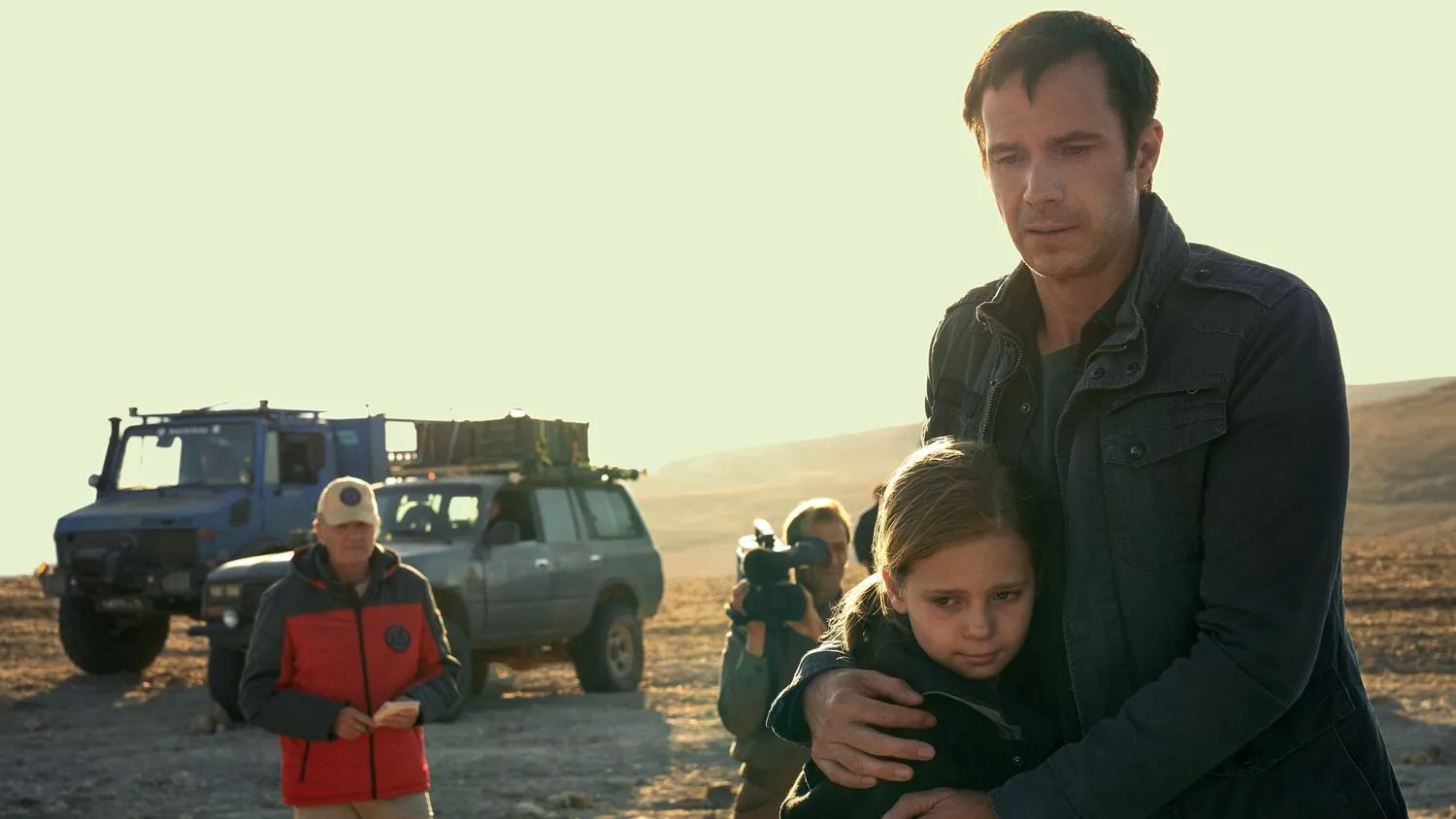 Mann, der ein junges Mädchen umarmt, während er in einer Wüste wartet. Im Hintergrund sind ein großer Lastwagen, ein Jeep, eine Person mit roter Jacke und Mütze sowie ein Mann mit einer großen Kamera zu sehen.