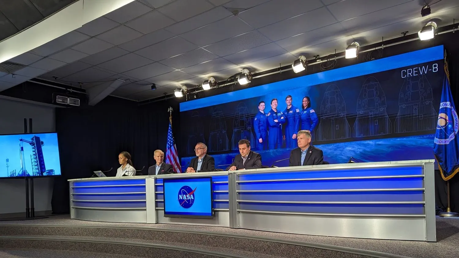 Menschen in Anzügen sitzen an einem Schreibtisch vor einem Poster mit vier Astronauten in Fluganzügen und dem Text Crew-8