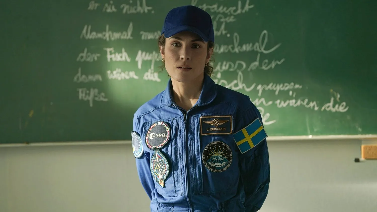 Eine Frau trägt einen blauen Astronautenoverall, der mit verschiedenen Aufnähern, darunter einer schwedischen Flagge, verziert ist. Außerdem trägt sie eine blaue Baseballmütze. Sie steht vor einer schwarzen Tafel, auf der mit Kreide etwas geschrieben steht.