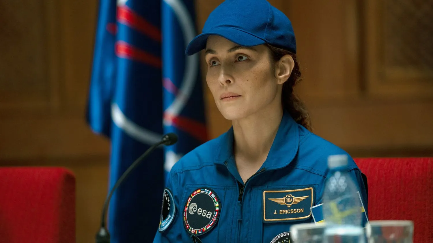 eine Frau in einer blauen Astronautenuniform sitzt an einem Schreibtisch