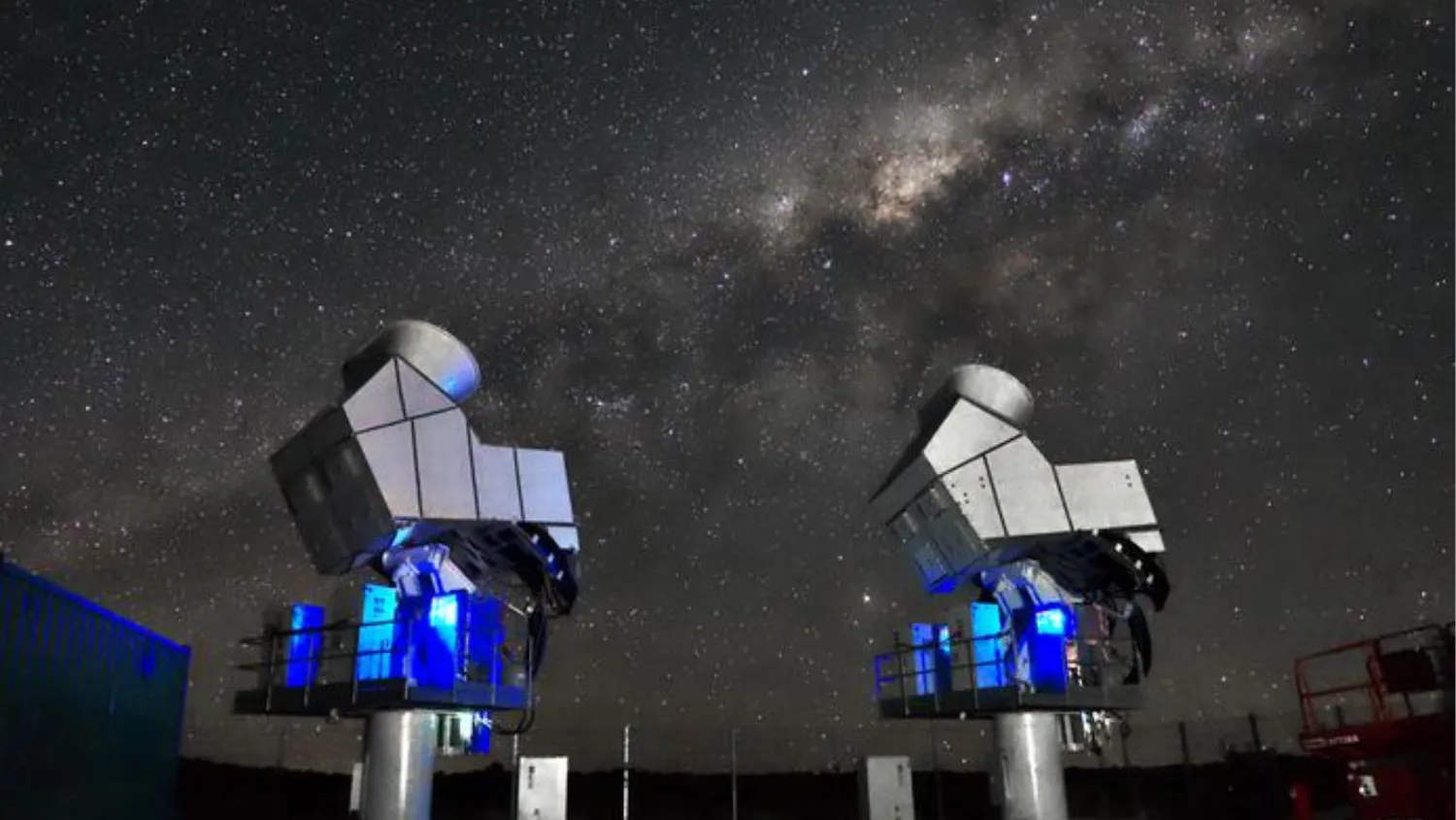 Zwei Metallgeräte mit blauen Lichtern an ihren Hälsen sind in den Himmel gerichtet. Am Himmel sind viele Sterne zu sehen und das Zentrum der Milchstraße ist sichtbar.