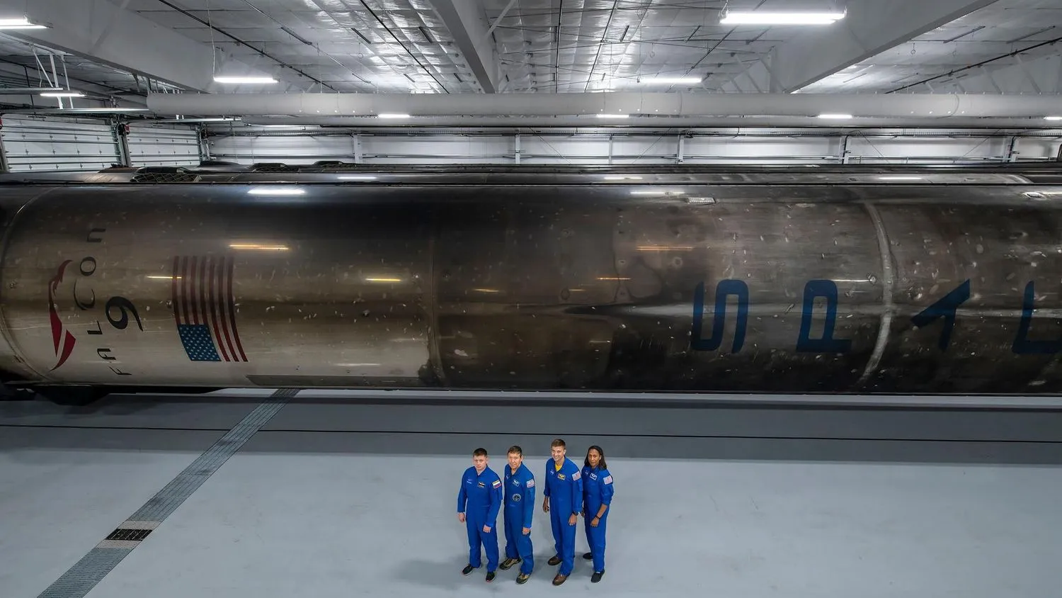 Vier Menschen in blauen Overalls stehen in einem großen weißen Hangar mit einer relativ niedrigen Decke. Hinter ihnen, auf der Seite liegend, befindet sich ein rußiger Raketenbooster, schwarz verschmiert von der Kohle, die bei früheren Flügen durch die Erdatmosphäre zurückkehrte.
