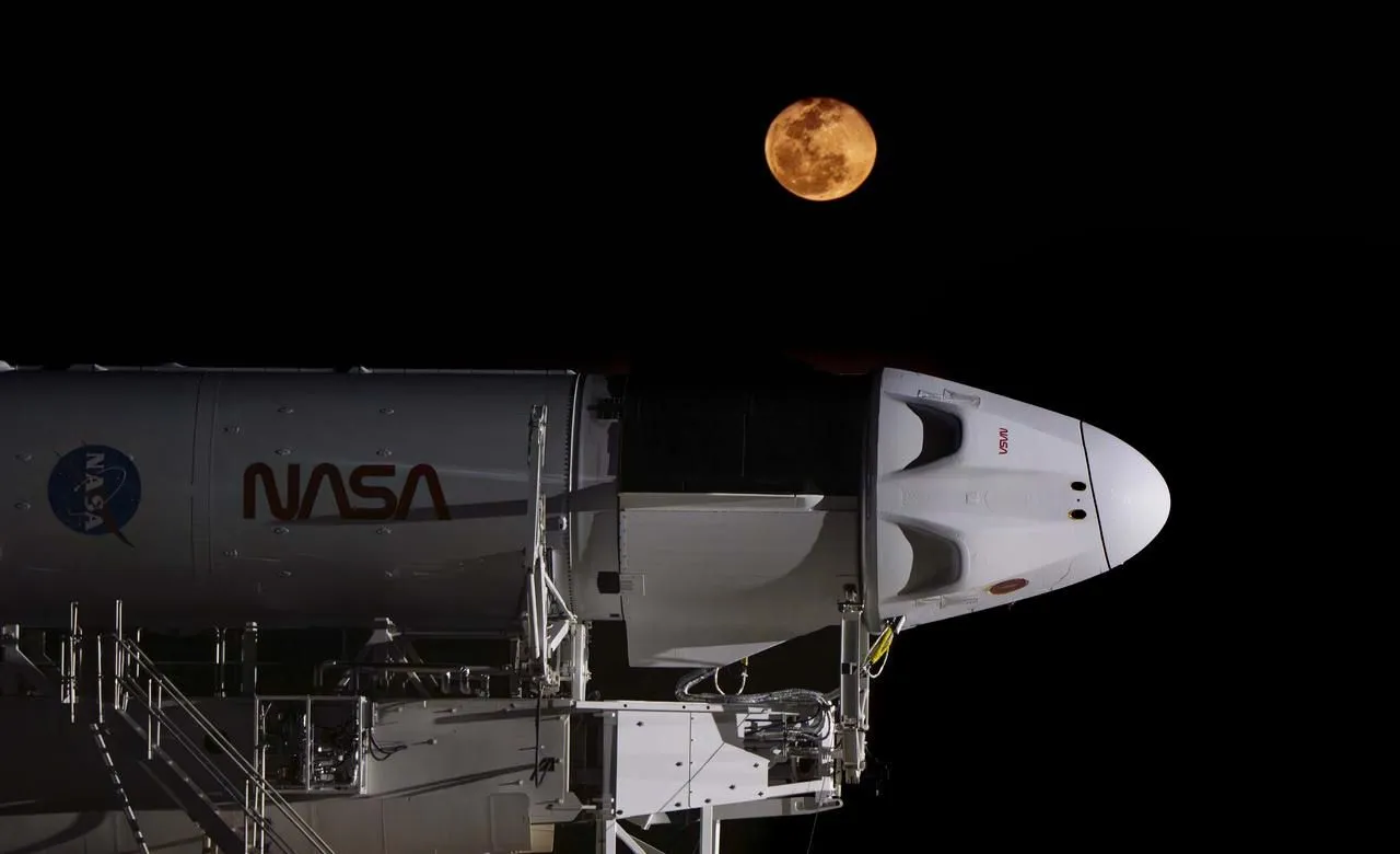 Man sieht den oberen Teil einer weißen Rakete, die in der Dunkelheit der Nacht auf Stützen liegt. Eine unsichtbare Lichtquelle leuchtet von der Raketenspitze herab. Ein orange-gelber Mond hängt am Himmel darüber.