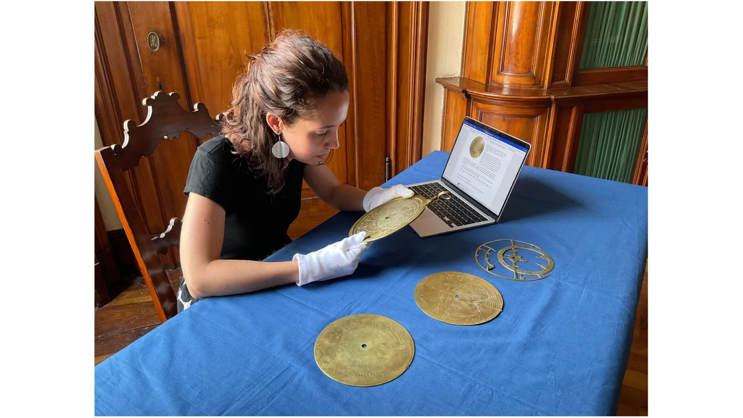eine Frau sitzt an einem Tisch und untersucht ein scheibenförmiges astronomisches Instrument aus Bronze