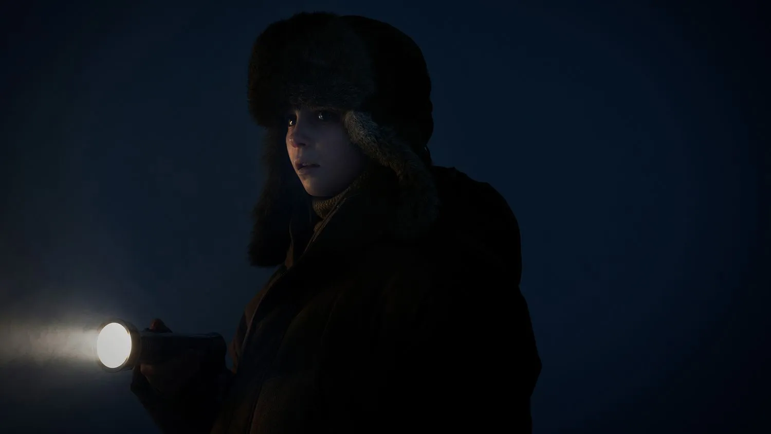 Junges Mädchen, warm eingepackt in Wintermütze und Mantel, hält eine Taschenlampe in der Hand, während sie im Dunkeln sucht.