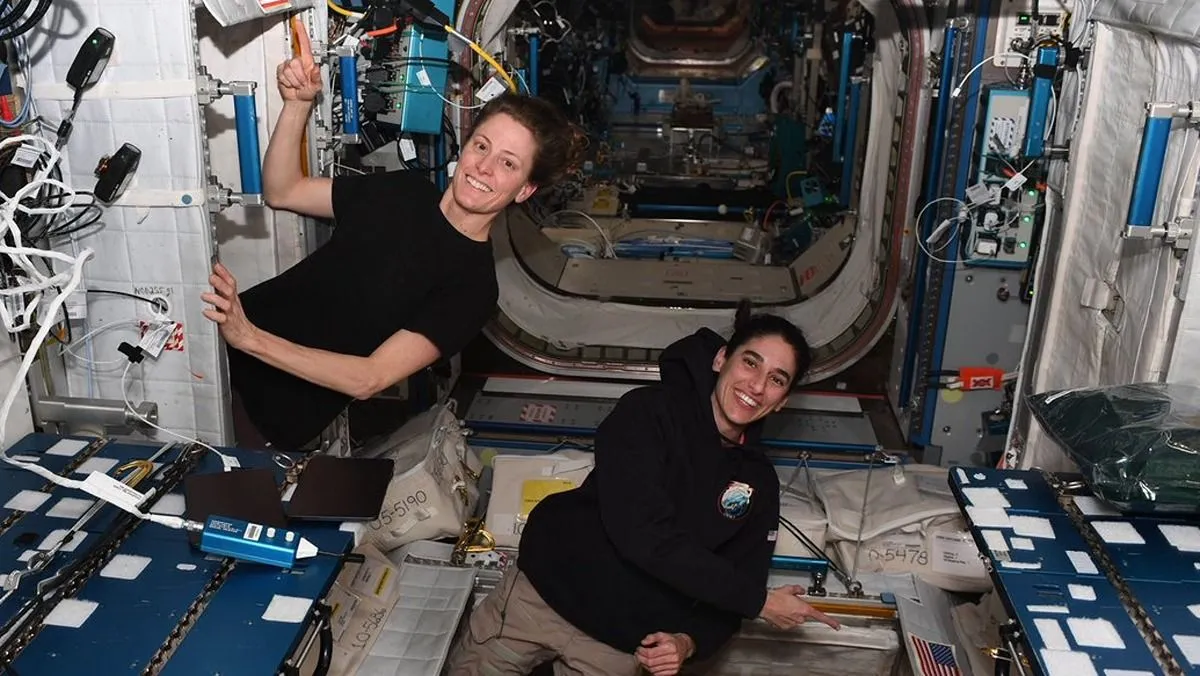 zwei astronautinnen schweben auf der internationalen raumstation. eine zeigt auf eine amerikanische flagge