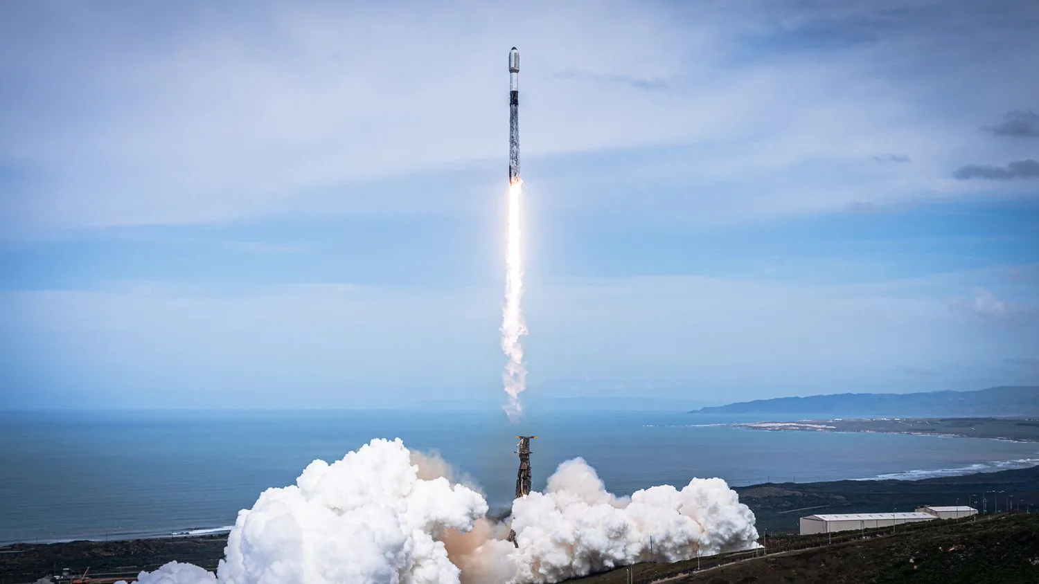 eine schwarz-weiße Spacex Falcon 9 Rakete startet in einen wolkenverhangenen blauen Himmel mit dem Meer im Hintergrund