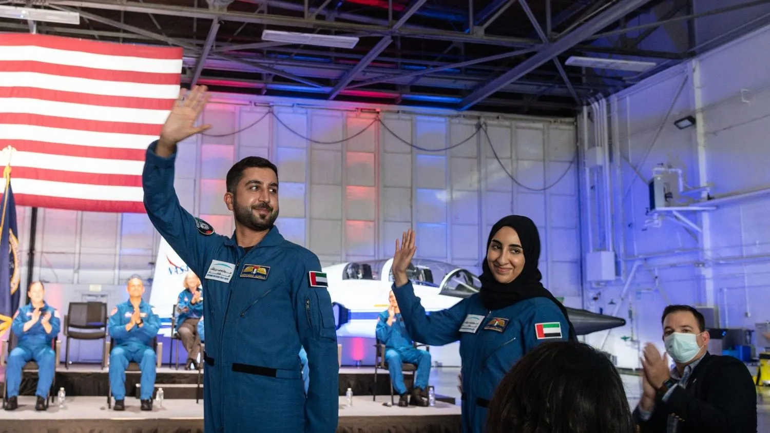 zwei astronautenkandidaten winken in fluganzügen vor einer us-flagge und einem t-38 jet. auf der bühne dahinter applaudieren sitzende astronautenkandidaten