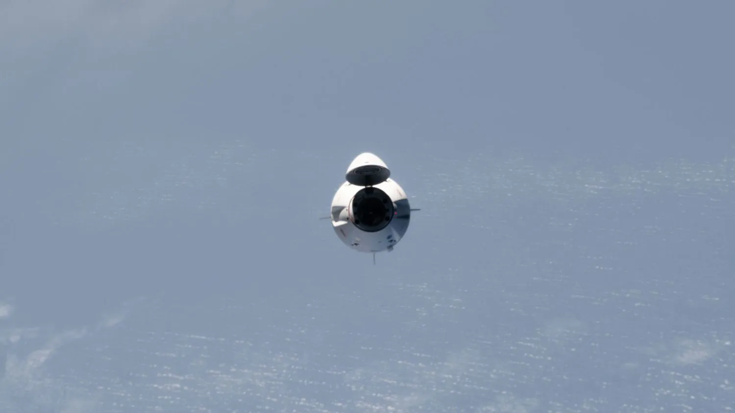eine weiße Raumkapsel nähert sich der internationalen Raumstation, mit ein paar Wolken über dem Ozean im Hintergrund