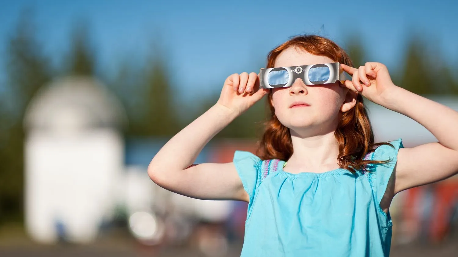 Ein Mädchen in einem blauen Oberteil hält eine Sonnenfinsternisbrille auf ihr Gesicht, während sie eine Sonnenfinsternis beobachtet.