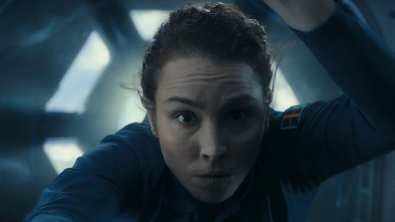 Nahaufnahme einer Frau in einem blauen Raumanzug (mit einem schwedischen Flaggenaufnäher am Arm), die in der Internationalen Raumstation schwebt. Sie blickt direkt in die Kamera.