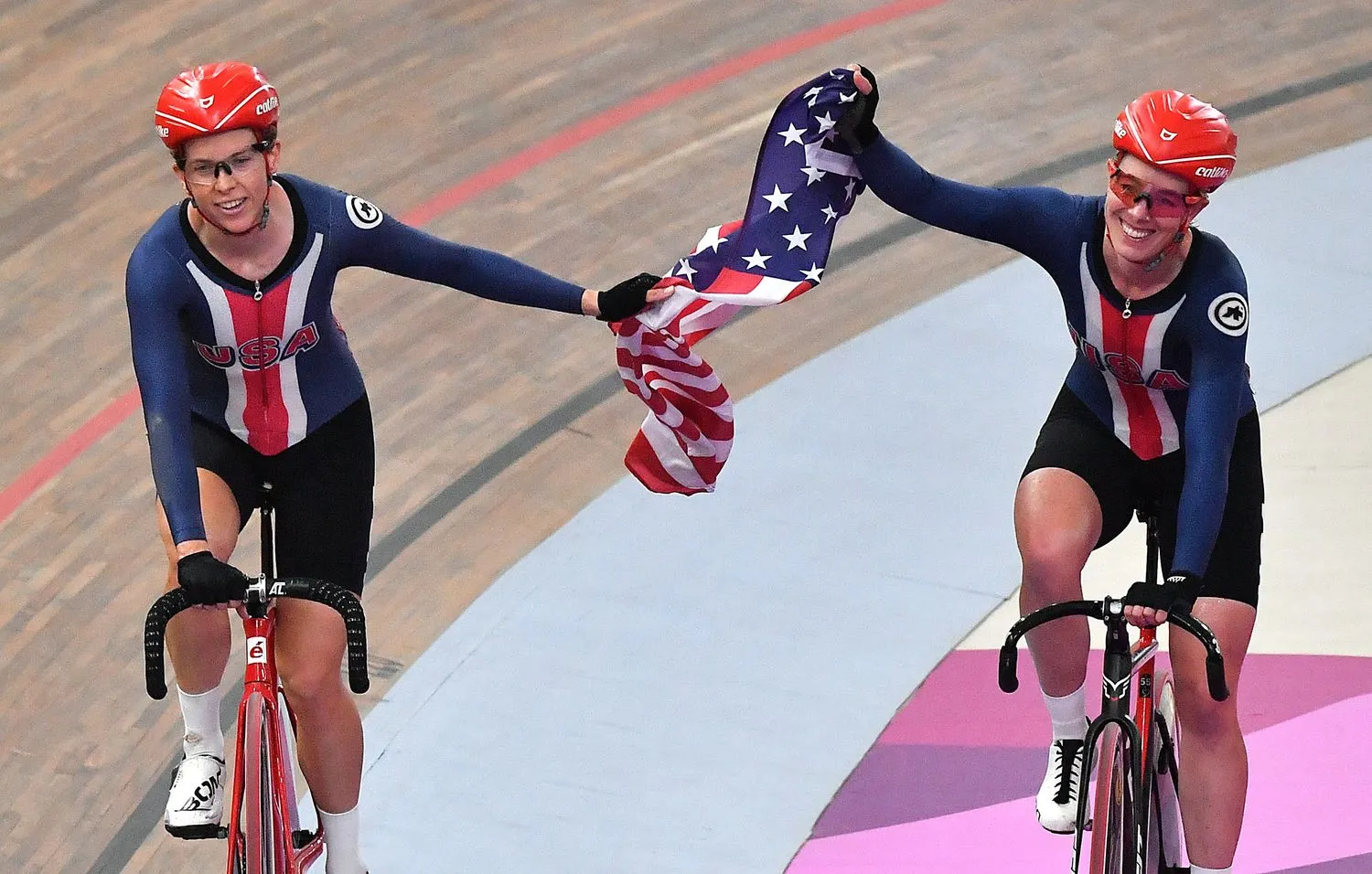 zwei Frauen auf einer Radrennbahn, auf Fahrrädern, die eine amerikanische Flagge halten